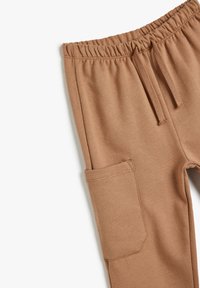 Pantalones jogger de color marrón claro con cintura elástica, cordón y un bolsillo lateral rectangular sobre un fondo blanco.