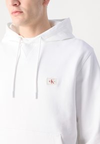 Biela bavlnená mikina s kapucňou na šnúrku, predná vrecko a obdĺžniková nášivka s logom v červenej a béžovej farbe. Hladká textúra, minimalistický dizajn.