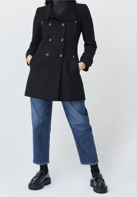 Cappotto nero doppiopetto con rever a punta, tessuto texturizzato e tasche laterali con zip. Abbinato a jeans blu a gamba dritta e scarpe con plateau nere.