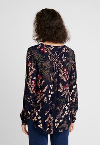 Blouse bleu marine à manches longues, motifs floraux et feuillages en rose, rouge et vert. Dotée d'un col rond et d'un détail froncé au dos.