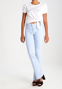 Light blue flared jeans with a high waist, paired with a white knotted t-shirt featuring black graphic text. Black heeled sandals.