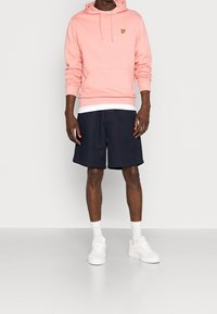 Felpa rosa chiaro con tasca frontale e logo, abbinata a pantaloni corti blu navy e sneakers bianche, con calzini bianchi alla caviglia. Design semplice e casual.
