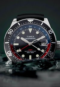 Timex Deepwater-horloge met een zwarte wijzerplaat, lichtgevende witte markers, rode GMT-accenten, roestvrijstalen lunette en zwarte rubberen band; waterbestendig tot 200 meter.