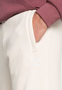 Světle krémové tepláky s měkkou texturou, které mají boční kapsy a malé bílé logo adidas na levém stehně.