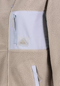 Beige fleece jacket with a light blue pocket, featuring a zip closure and a rubber logo patch. Soft texture and contrasting materials.