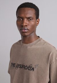 Camiseta oversized marrón con cuello redondo y diseño de texto en negro, que presenta acentos de tachuelas plateadas para añadir textura.