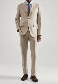 Beige pak met een jasje met twee knopen, notch revers en rechte broekspijpen. Gecombineerd met een lichtblauwe overhemd en bruine instappers.