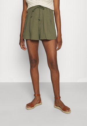 Femme portant un short olive à cordon de serrage et des sandales marron à bride de cheville, se tenant devant un fond blanc uni.