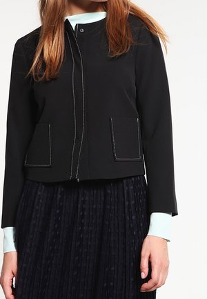 Kvinde iført en sort cropped jakke med hvide syninger over en lyseblå bluse med lange ærmer og en sort plisseret nederdel med prikkemønster.