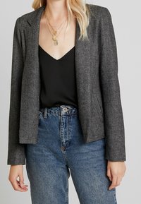Grauer Fischgrätblazer mit strukturiertem Design, Reverskragen und langen Ärmeln, getragen über einem schwarzen Oberteil und hochtaillierten Jeans.
