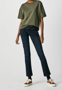 Person som bär en olivgrön t-shirt med kort ärm, mörkblå jeans, svarta strumpor och bruna och vita sneakers står framför en enfärgad bakgrund.