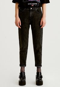 Person som bär svarta tapered jeans med uppvikt mudd, fiskenätstrumpor och svarta plattformsskor med spännen, stående mot en enfärgad bakgrund.