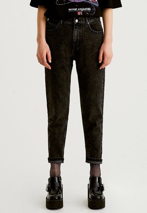 Personne portant un jean noir ajusté avec des ourlets retroussés, des chaussettes en résille et des chaussures à plateforme noires avec des boucles, se tenant debout contre un fond uni.
