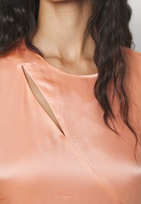 Persikofärgad satinblus med en rund halsringning och en diagonal slits, texturerad tyg och en mjuk finish. Subtil lyster förbättrar det övergripande utseendet.