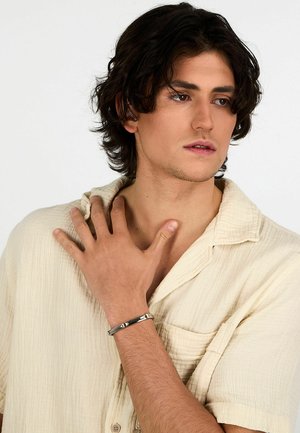 Bracciale in metallo argenteo con accenti geometrici indossato su una mano. Modello in una camicia bottonata beige chiaro e testurizzata, mano appoggiata vicino al collo.