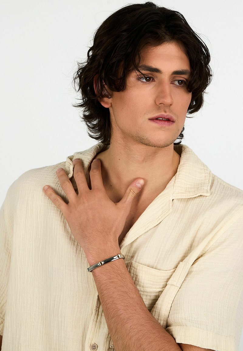 Bracciale in metallo argenteo con accenti geometrici indossato su una mano. Modello in una camicia bottonata beige chiaro e testurizzata, mano appoggiata vicino al collo.
