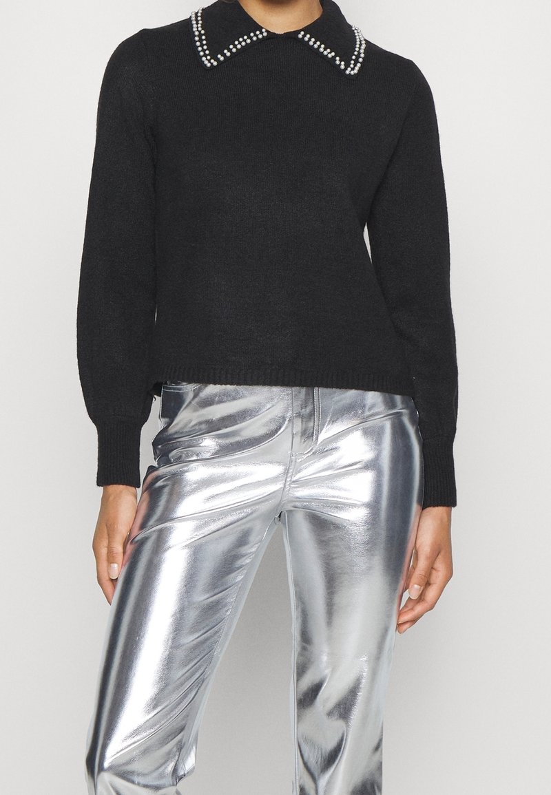 Personne portant un pull noir avec un col orné de perles et un pantalon argenté métallique brillant, debout avec les bras détendus le long du corps.