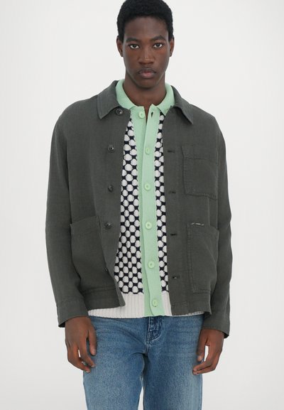 Veste verte foncée, fermeture à boutons, deux poches avant, portée sur une chemise légère verte à motif pois noir et blanc, accompagnée d'un jean bleu.
