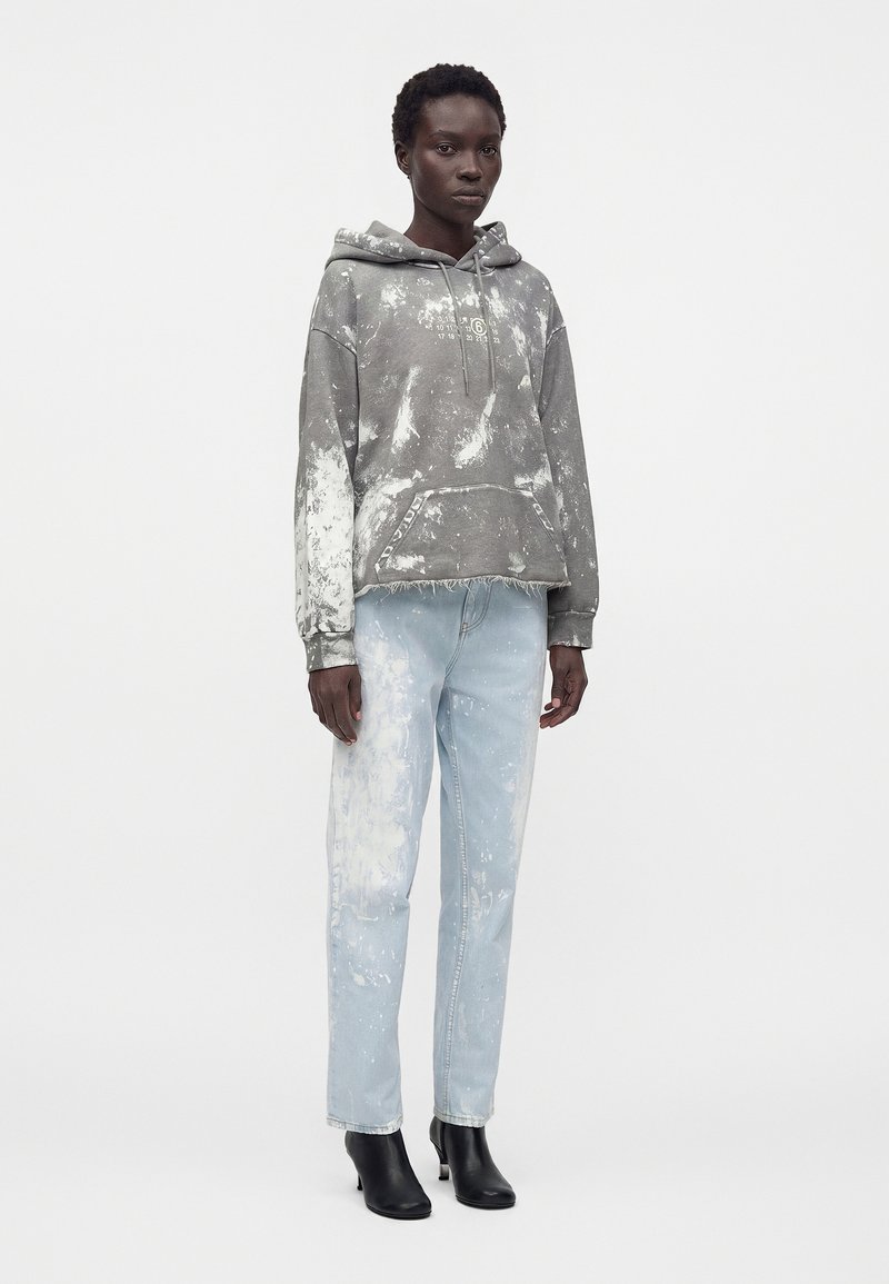 Standing person wearing a gray hooded sweatshirt with white splatter and light blue splattered jeans, paired with black ankle boots.