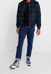 Giacca bomber a quadri in blu navy, verde e nero, con polsini e orlo a coste. Abbinata a jeans in denim con orlo arrotolato e sneaker bianche con dettagli blu.