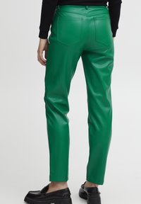 Persona vestida con pantalones de cuero verde brillante y mocasines negros, de pie con las manos relajadas a los lados, vista desde la espalda.