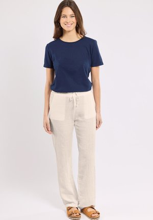 Femme souriante, vêtue d'un haut bleu marine à manches courtes, d'un pantalon beige à cordon de serrage et de sandales marron clair, se tenant debout devant un fond clair uni.