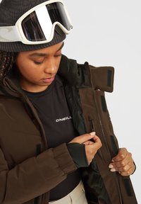 Bruine geïsoleerde jas met een hoge kraag, zijritsen en klittebandaccenten. Draag het over een zwart shirt, vergezeld van witte goggles.