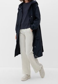 Tummansininen vedenpitävä takki, jossa on huppu, napit ja sivutaskut, yhdistettynä tummaan toppiin ja vaaleisiin leveisiin housuihin.