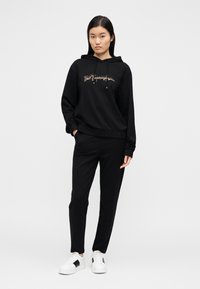 Μαύρο φούτερ με κουκούλα και γραφικό σχέδιο, σε συνδυασμό με μαύρο παντελόνι και λευκά αθλητικά με μαύρες λεπτομέρειες. Ομαλό, casual ύφασμα.