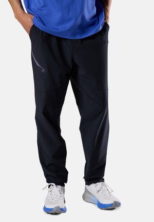 Mann trägt schwarze Sporthose mit Reißverschlusstasche und weiße Laufschuhe mit blauen Sohlen, Hände in den Taschen, vor einfachem Hintergrund.