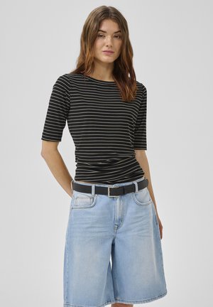 Kobieta w czarnej bluzce w paski z krótkim rękawem, włożonej do jasnoniebieskich, dżinsowych szortów do kolan z czarnym paskiem, stojąca na jednolitym tle.