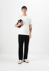Ung mann med kort hår, iført hvit t-skjorte, svarte bukser, hvite joggesko, og holder en svart clutch-veske mot en hvit bakgrunn.