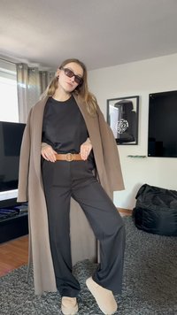 Persoană tânără purtând ochelari de soare, jachetă maro, cămașă și pantaloni negri cu o curea maro, pantofi bej, stând în un living.