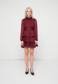 Burgundské šaty s více vrstvami, dlouhými rukávy, texturovanou látkou, nařaseným pasem a vroubkovaným spodním lemem. Nosit s černými lodičkami se špičatou špičkou.