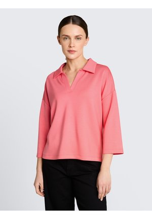 Femme portant un haut ample rose avec un col et un décolleté en V, associé à un pantalon noir, debout devant un fond clair uni.