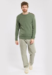 Zielony sweter prążkowany z guzikami na ramionach, połączony z jasnobeżowymi spodniami cargo i białymi sneakersami. Gładkie tło, widok całej postaci.