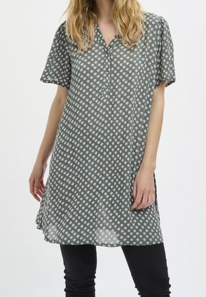 Woman wearing a green flowy tunic with white floral print, short sleeves, and black pants, standing against a plain white background.