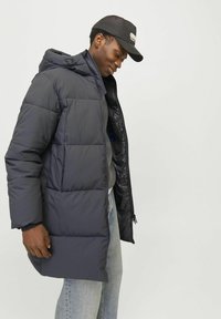 Mørkeblå pufferjakke med hætte, der har et kviltet design, lynlås lukning og sidelommer. Bæres over en grå sweater og jeans.