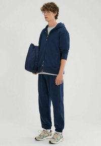 Felpa zip-up blu marina con una maglietta grigia sotto, abbinata a pantaloni jogger coordinati e sneakers chiare con dettagli verdi.