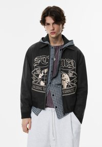 Musta takki, jossa on beige maapallokuvio, päällystettynä ruudullisen paidan päällä. Malli käyttää harmaita collegehousuja. Rakenteellinen kangas, rento istuvuus.