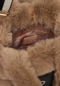 Fodera in pelliccia sintetica marrone con un interno in tessuto beige liscio. Presenta un'etichetta del logo cucita e un dettaglio con bottone dorato.