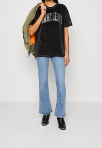 Persona que lleva una camiseta negra de "Tommy Jeans", jeans acampanados de color azul claro, botas negras, y sostiene una chaqueta verde oliva con forro naranja sobre un hombro.