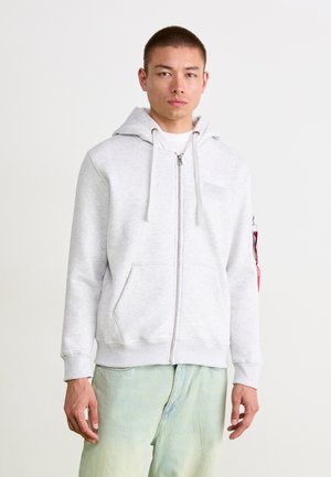 Veste à zip gris clair avec une poche kangourou, des cordons et un patch sur la poitrine. Dispose d'un accent rouge sur la manche. Coupe décontractée.
