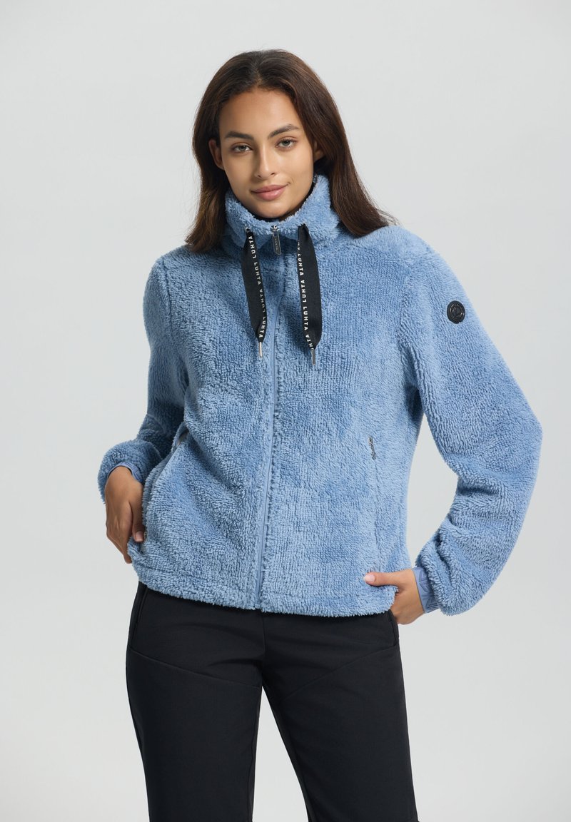 Frau trägt eine hellblaue, flauschige Zip-Jacke mit schwarzen Kordeln und schwarze Hosen, Hände in den Jacken Taschen, neutraler Hintergrund.