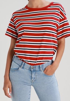 Personne portant une chemise à manches courtes rayée horizontalement rouge, blanche et noire rentrée dans un jean bleu clair, une main dans la poche.