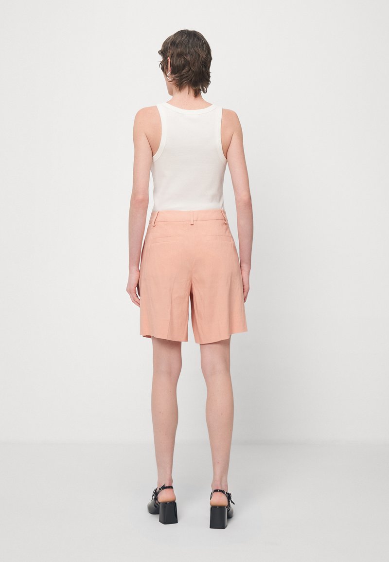 Frau, die mit dem Rücken zur Kamera steht, trägt ein weißes ärmelloses Oberteil, pfirsichfarbene knielange Shorts und schwarze Absatzsandalen vor einem einfarbigen Hintergrund.