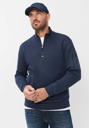 Uomo che indossa una felpa blu navy con zip parziale e tasca con zip sulla manica, cappellino blu navy abbinato e jeans blu, che guarda leggermente verso l'alto.