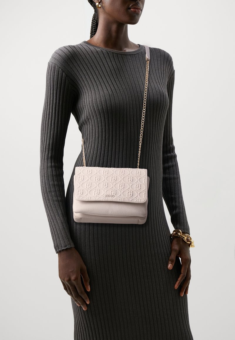 Kobieta w ciemnej, prążkowanej sukience, niesie jasnoróżową, teksturowaną torebkę na pasku łańcuszkowym i nosi złotą bransoletkę na nadgarstku.