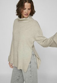 Pull à col roulé beige en tricot doux, avec des fentes sur les côtés, des manches longues et une coupe décontractée, associé à un jean taille haute gris.