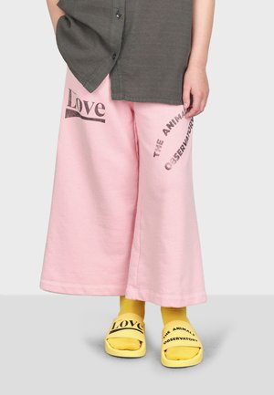 Rosa weite Sweatpants mit dem Aufdruck "Love" und "THE ANIMALS OBSERVATORY." Kombiniert mit gelben Slide-Sandalen, die kontrastierenden schwarzen Text aufweisen.